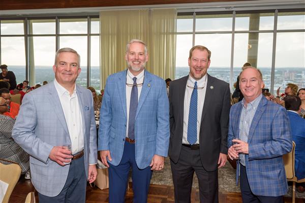 Four men in suits smile at the camera. The men on either end are holding drinks. The two in the middle have glasses hung around their next. The smile and laugh pleasantly.